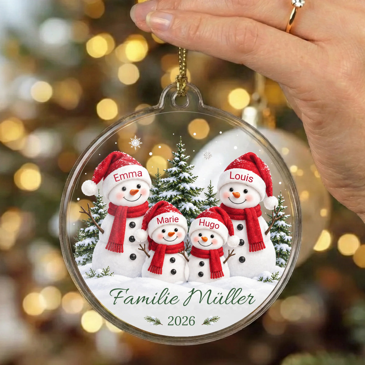 Personalisierter Acrylglas-Christbaumschmuck Schneemannfamilie mit vier Namen, Wunschtext und Jahr in der Hand vor warmen Weihnachtslichtern
